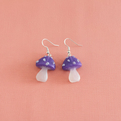 Mushroom-shaped earrings with purple caps and white bases on a pink background