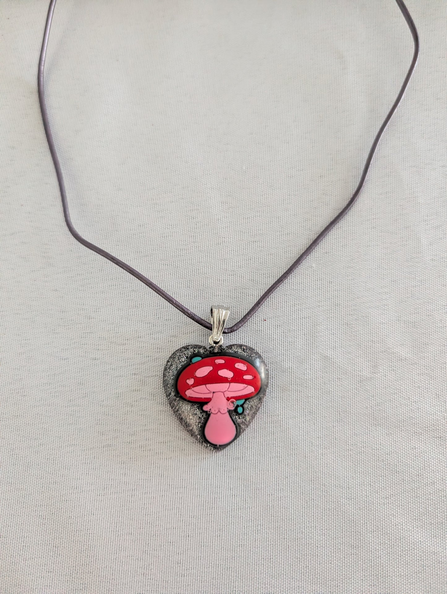 Heart-shaped pendant with a red mushroom design on a purple cord against a light gray background