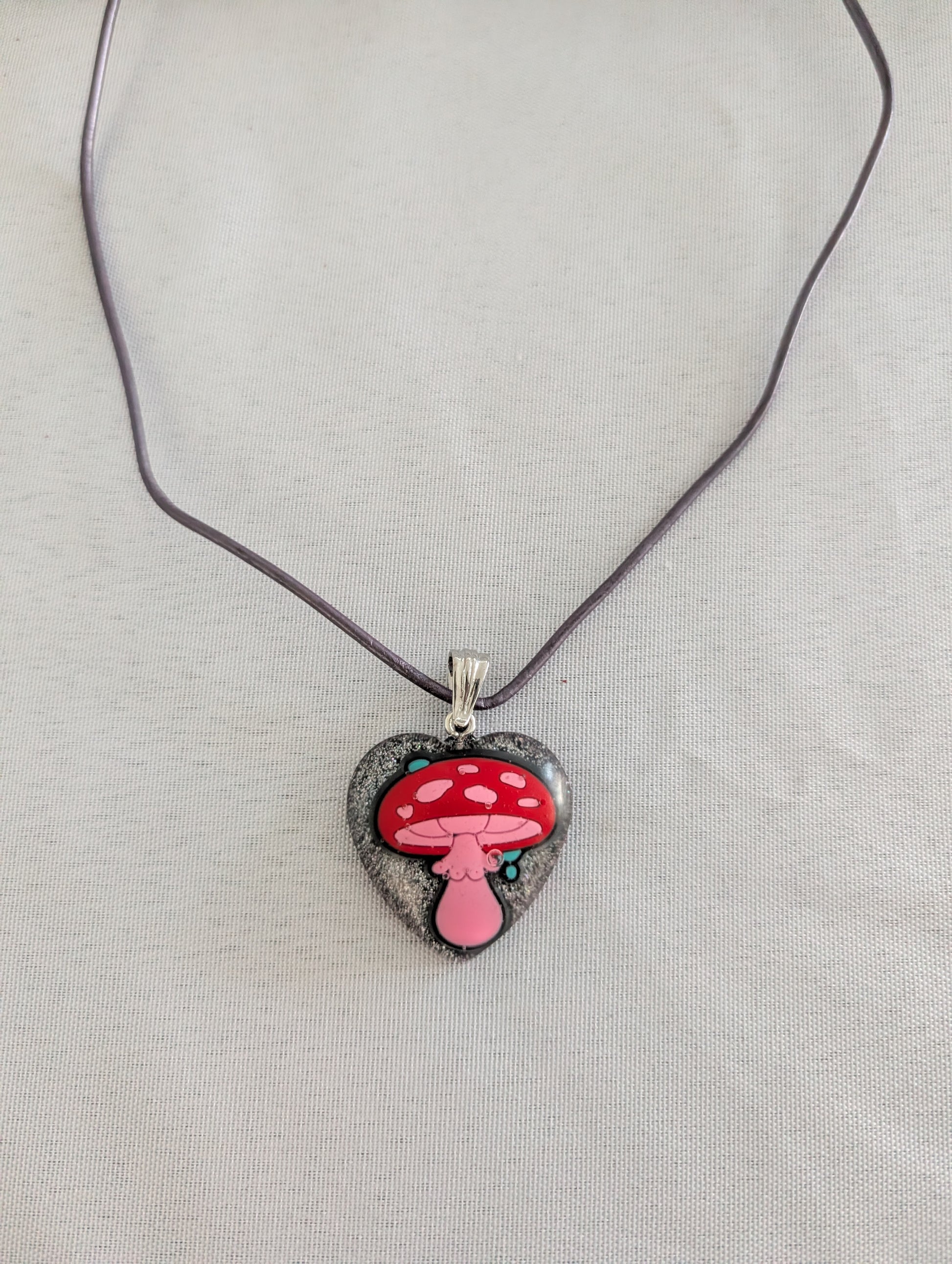 Heart-shaped pendant with a red mushroom design on a purple cord against a light gray background