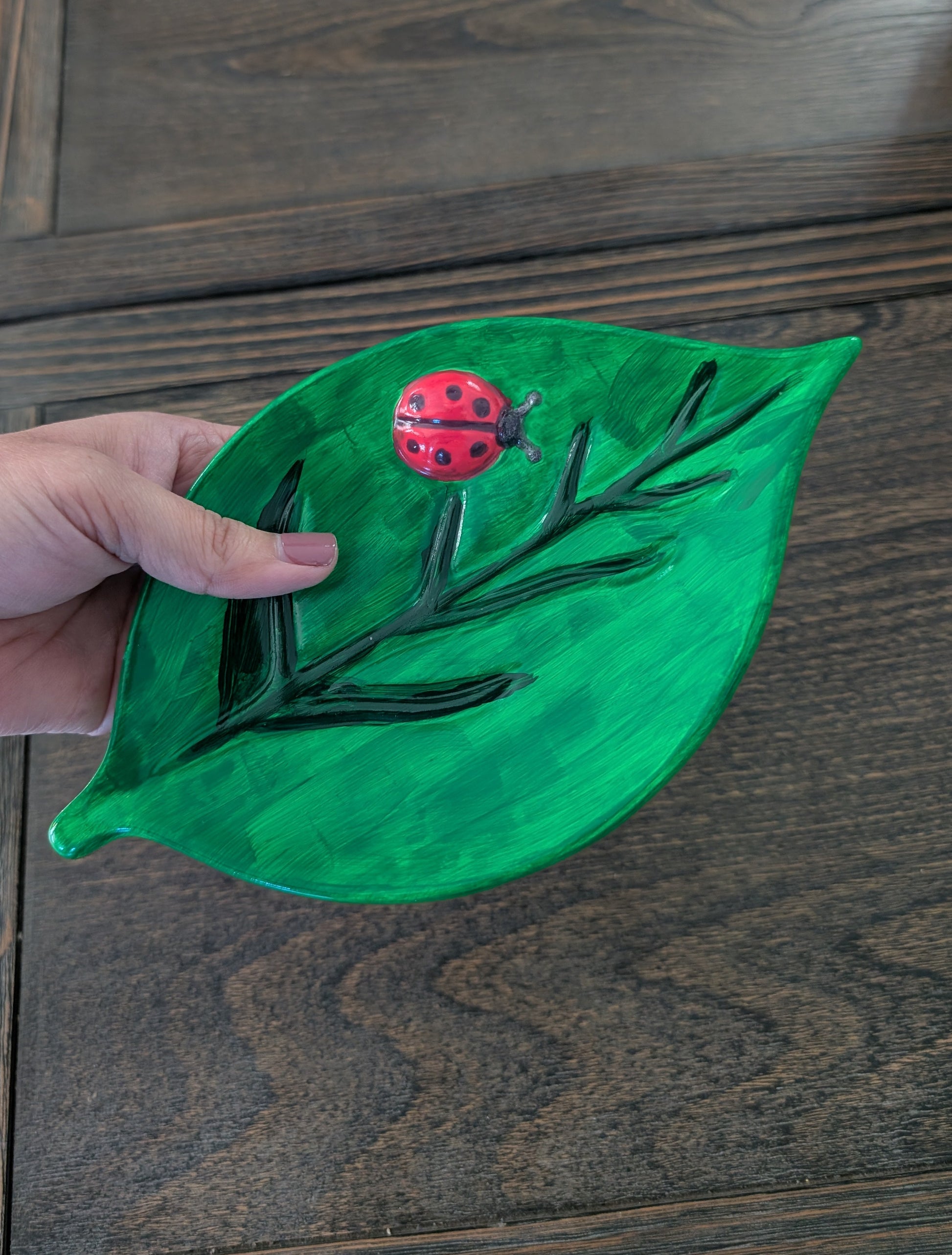 Hand holding a green leaf-shaped object with a red ladybug on a wooden surface