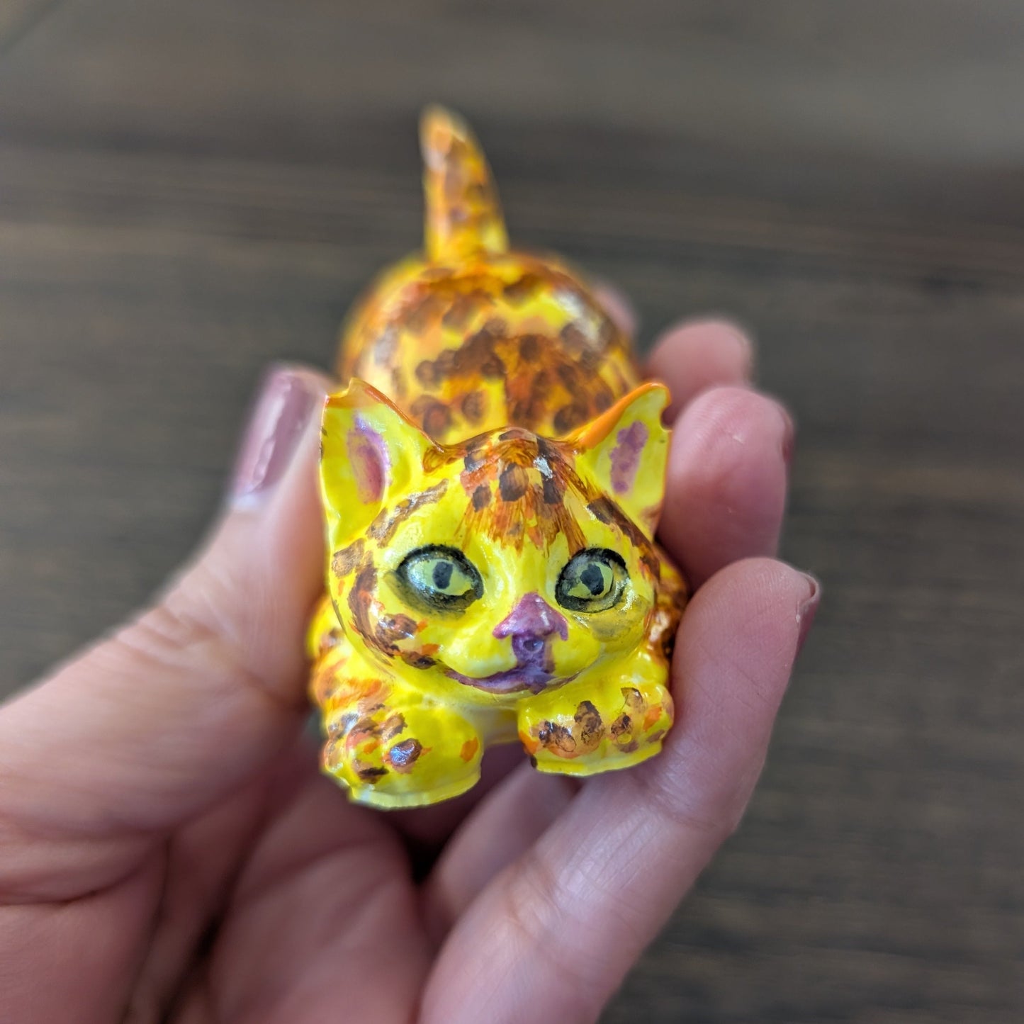 Hand holding a small figurine of a cat with yellow and orange pattern on a wooden surface