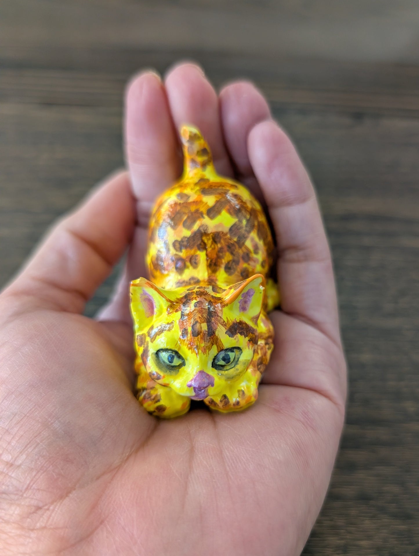 Hand holding a small figurine of a yellow cat with green eyes on a wooden surface.