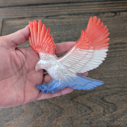 Hand holding a resin eagle figurine with red and blue wings on a wooden surface