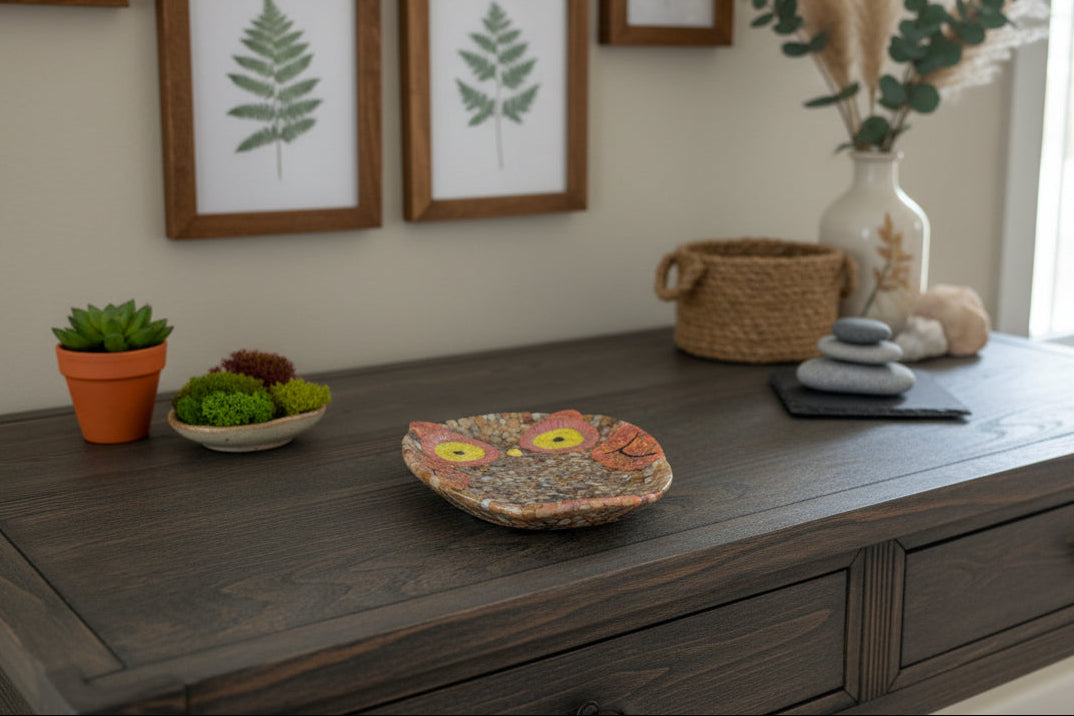 Decorative owl plate on a wooden surface with plants and framed artwork in the background.