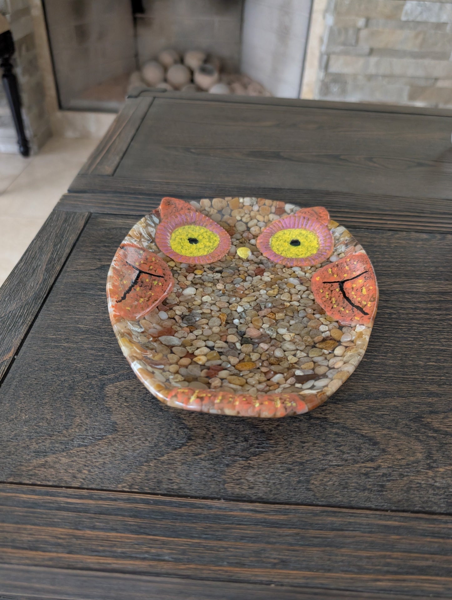 Decorative owl plate made of pebbles on a wooden surface with a fireplace in the background.