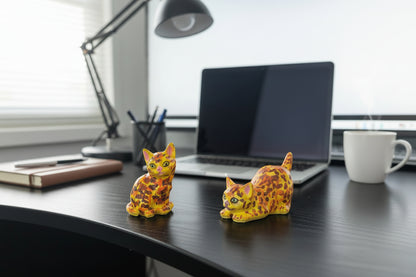 Two colorful cat figurines on a dark surface with a laptop in the background.