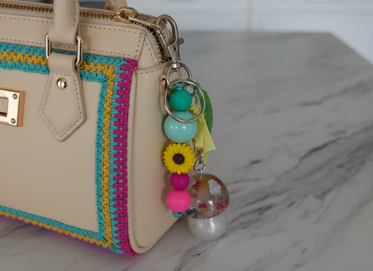 Colorful keychain with beads and a sunflower charm attached to a beige handbag on a marble surface.