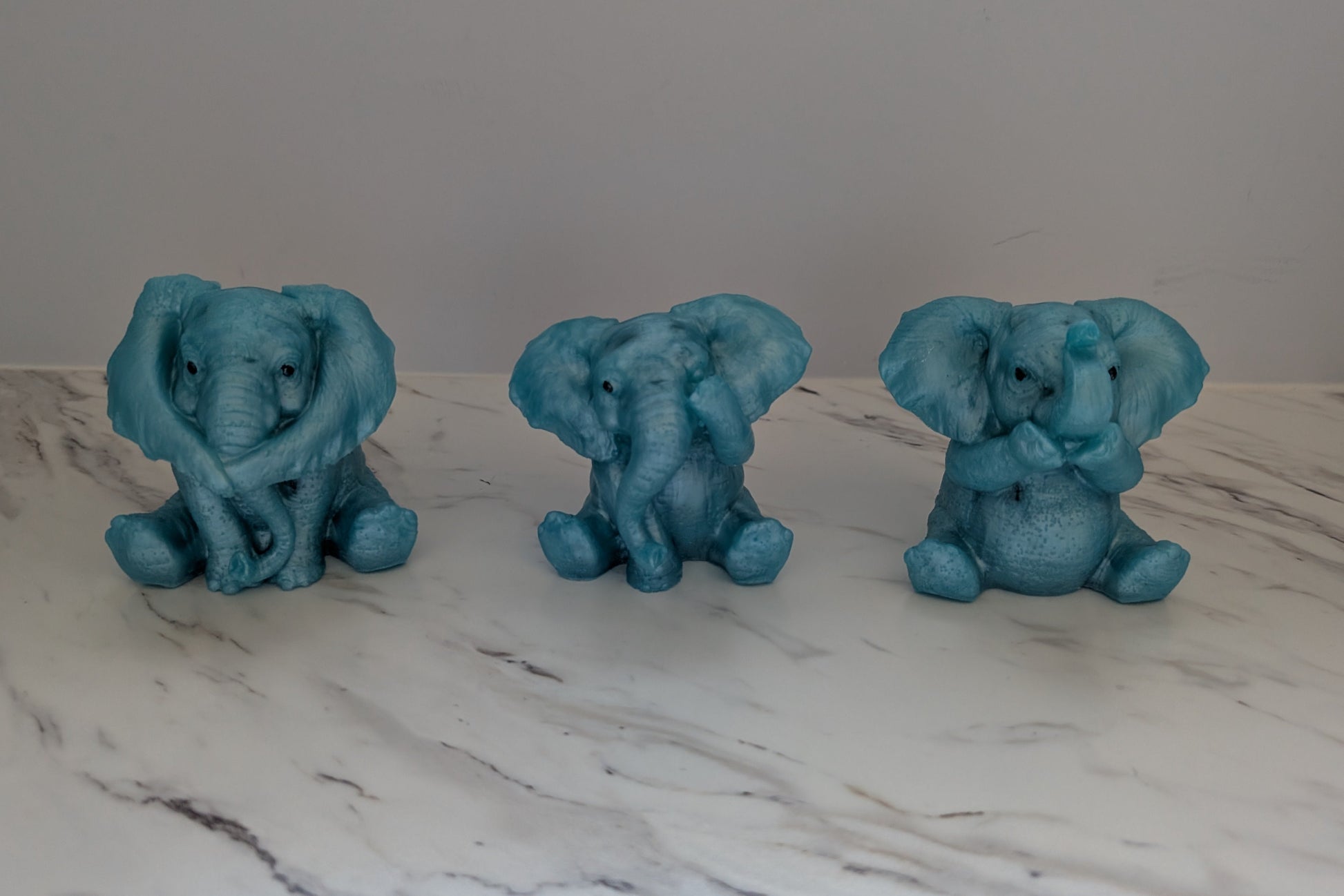 Handmade set of three blue resin baby elephant figurines representing see no evil, hear no evil, and say no evil on a marble surface.