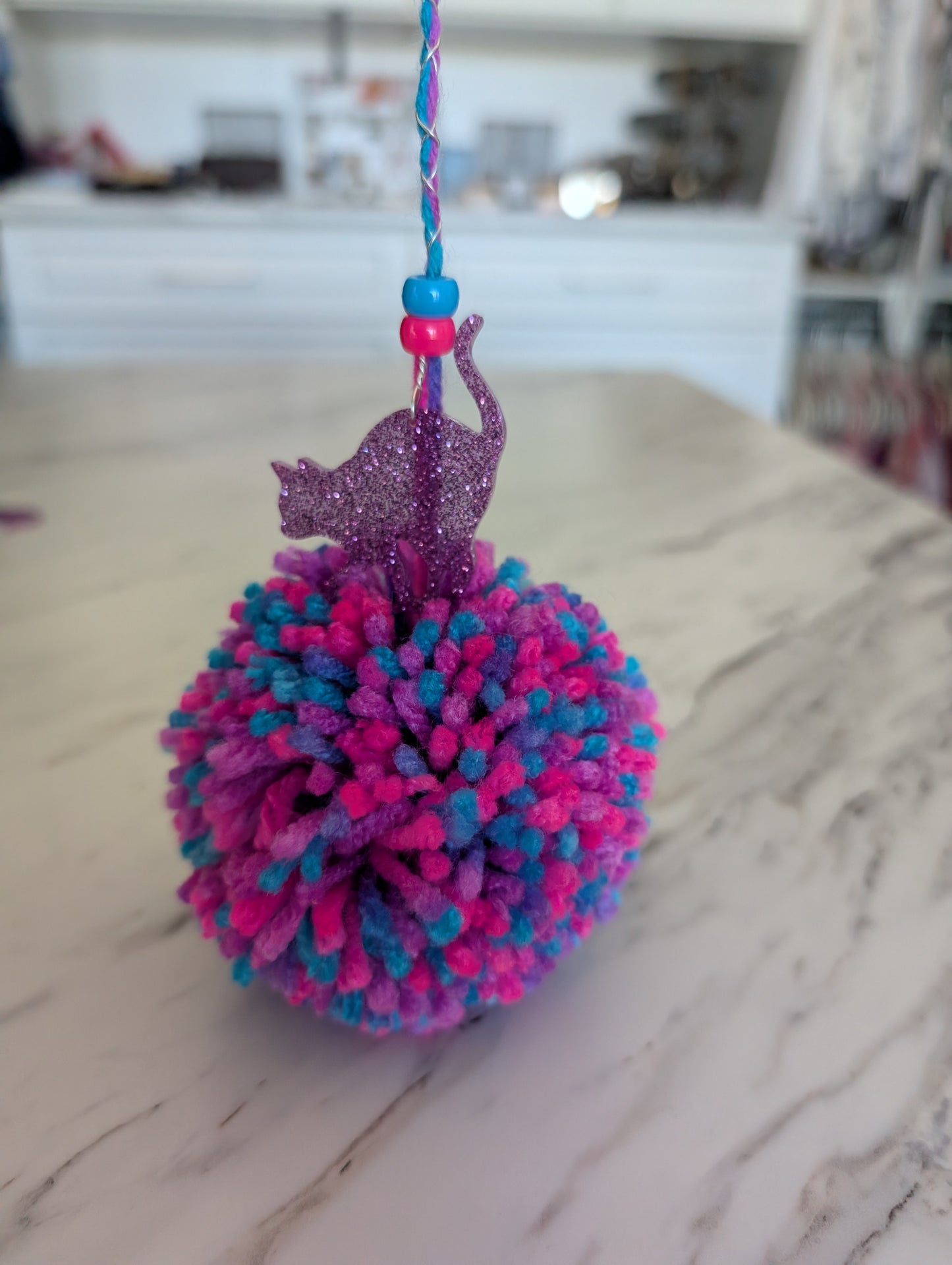 Pink and blue pom-pom with a cat charm on a marble surface