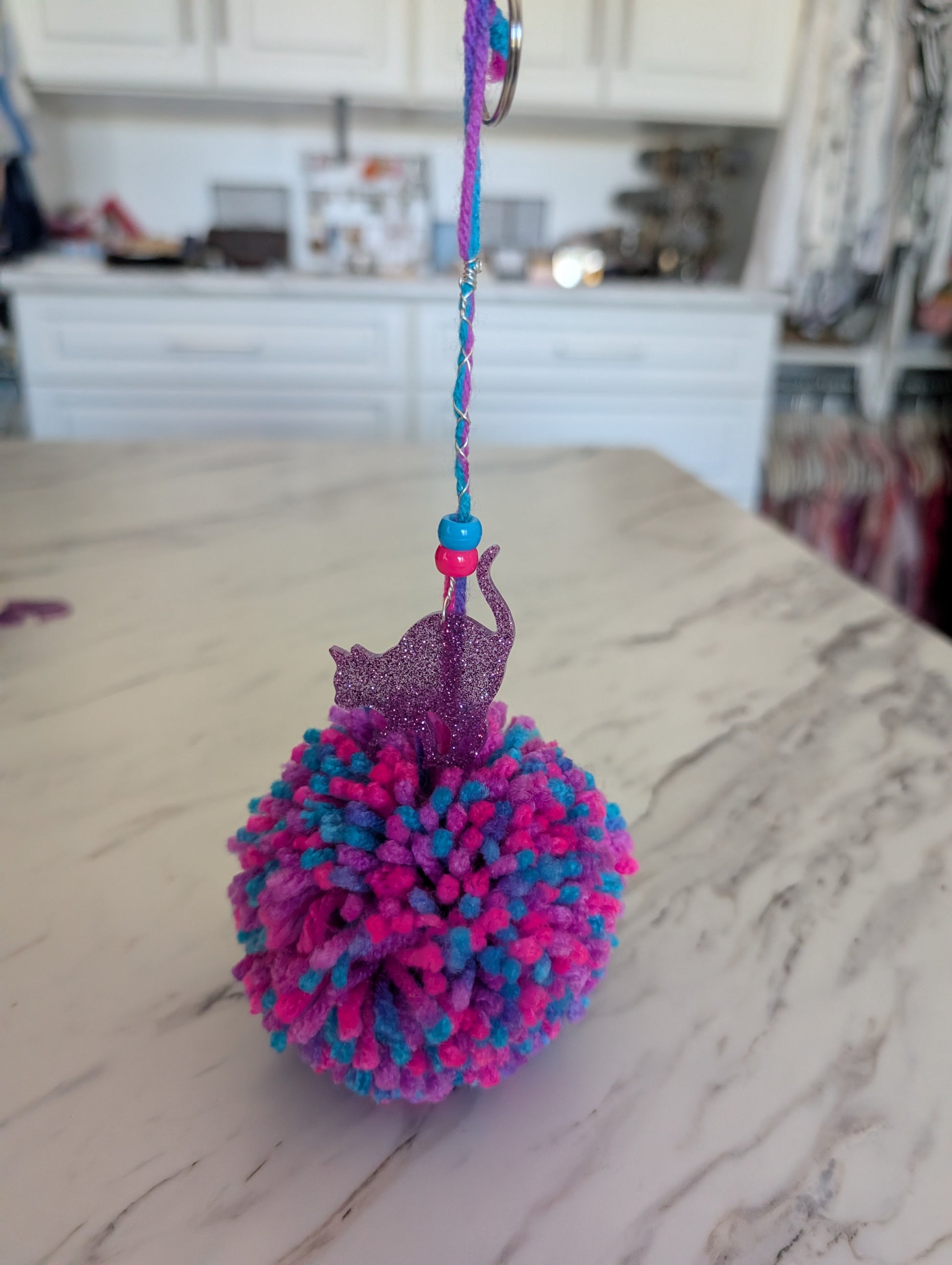 Colorful pom-pom keychain with a cat charm on a marble countertop.