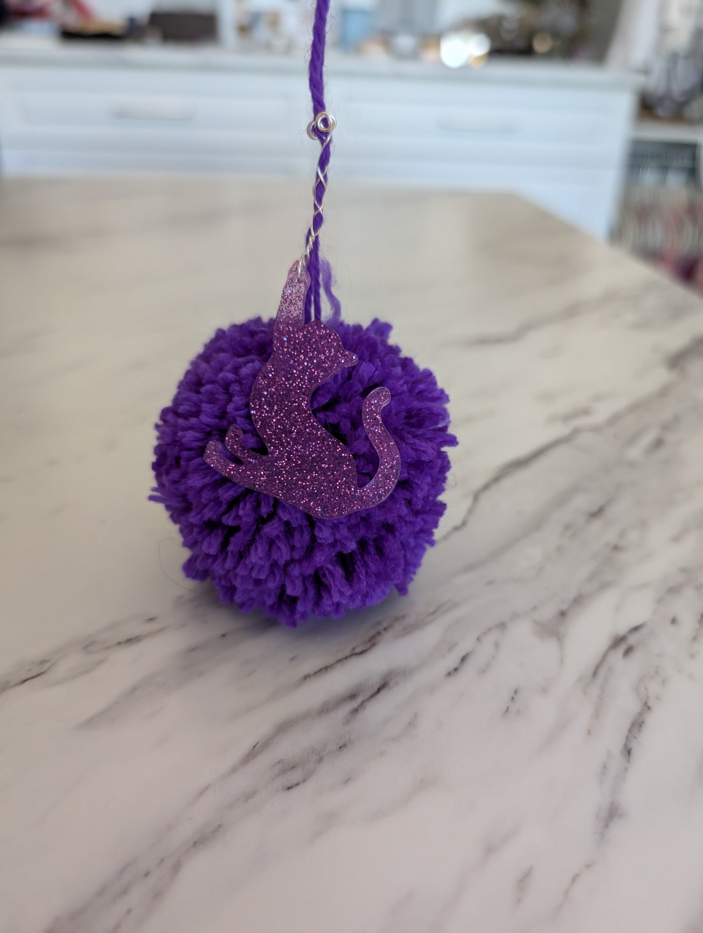 Purple pom-pom ornament with a glittery cat design on a marble counter top.
