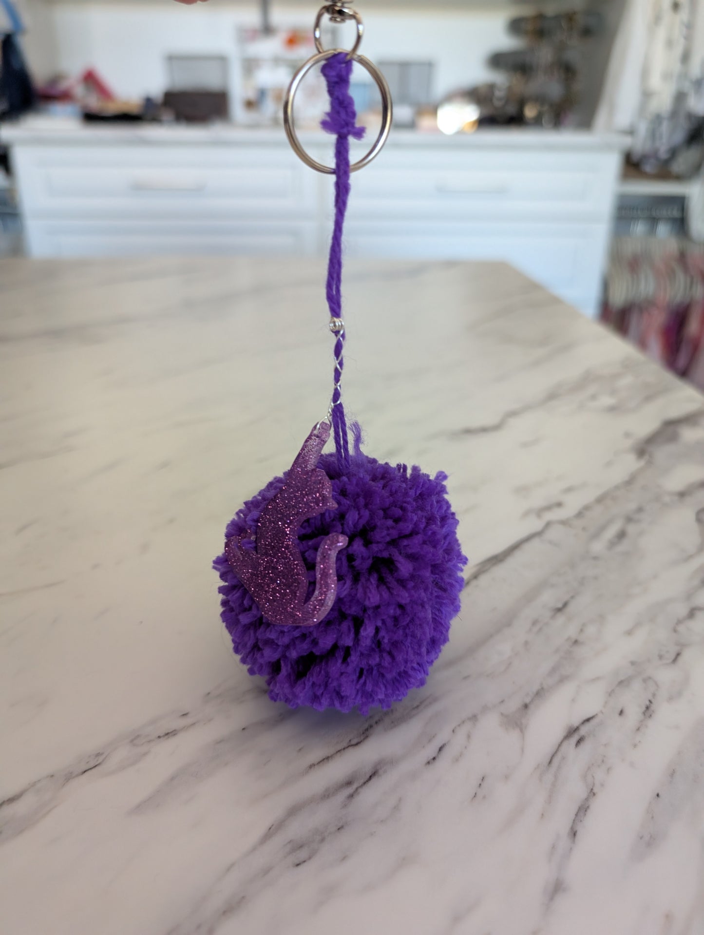Purple pom-pom keychain with a cat design on a marble surface