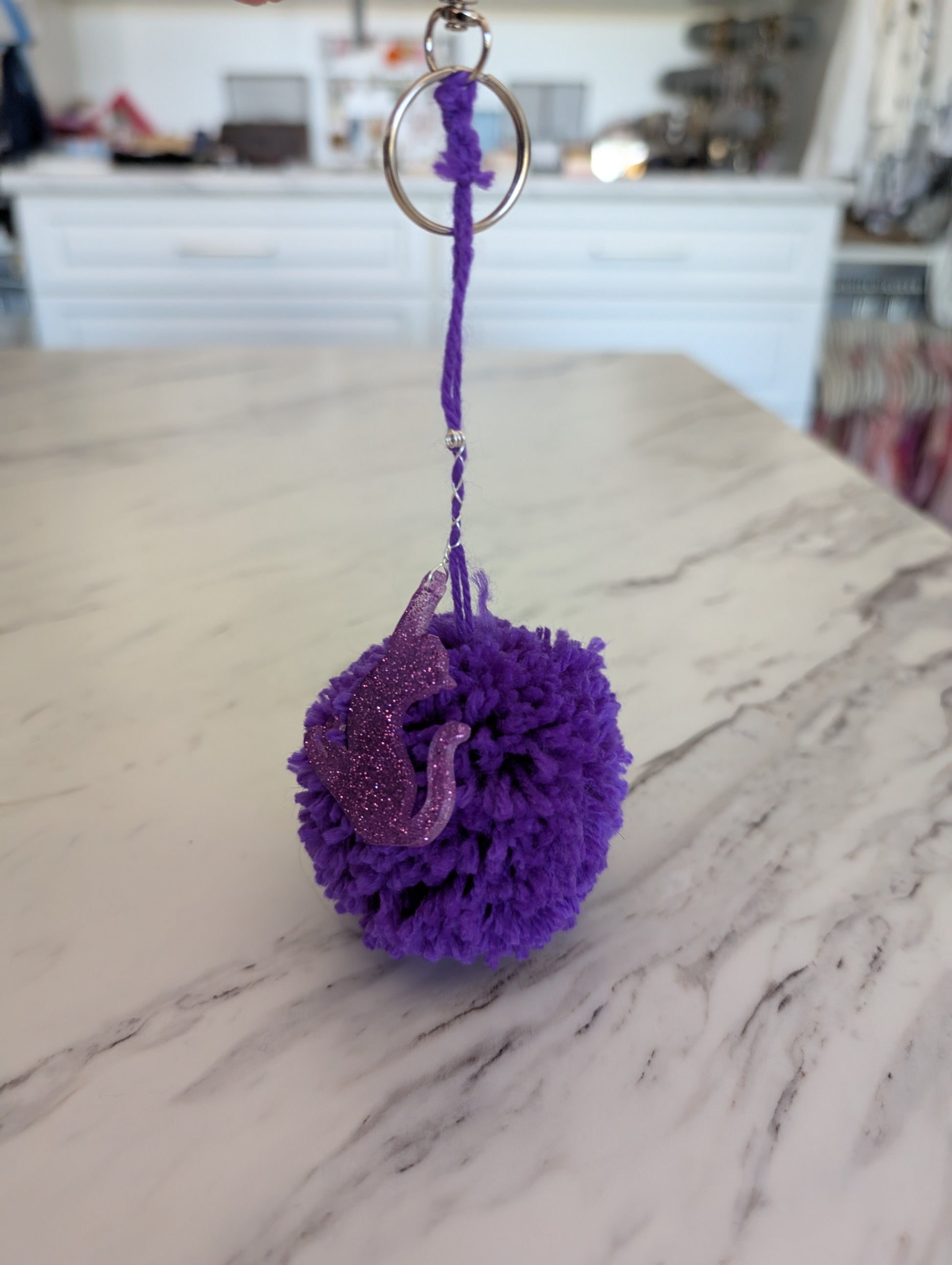Purple pom-pom keychain with a cat design on a marble surface