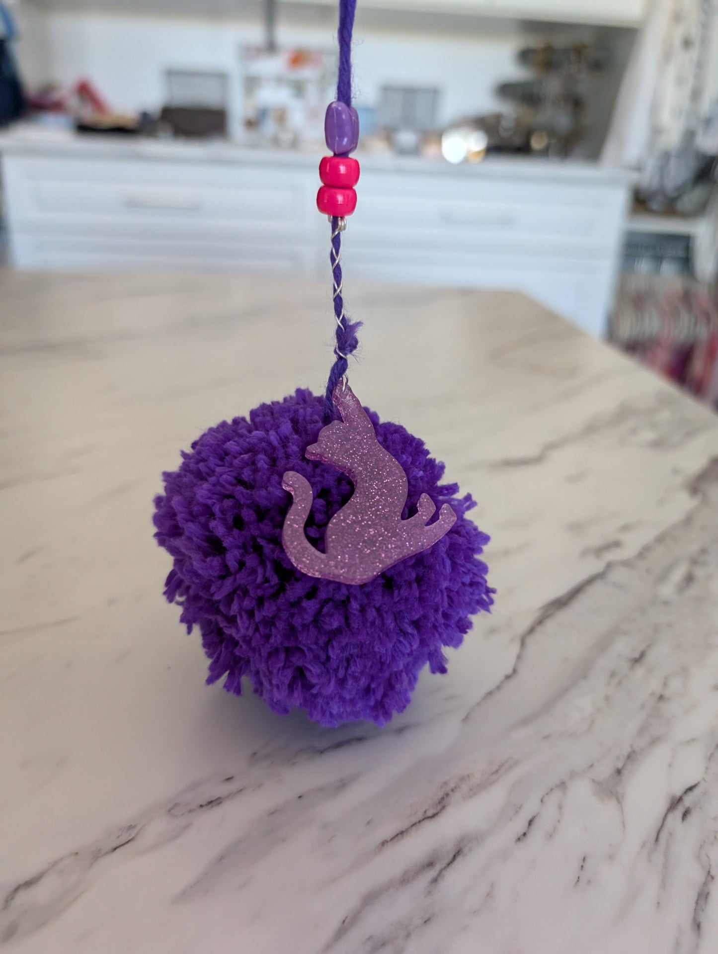 Purple pom-pom charm with a cat charm on a marble surface
