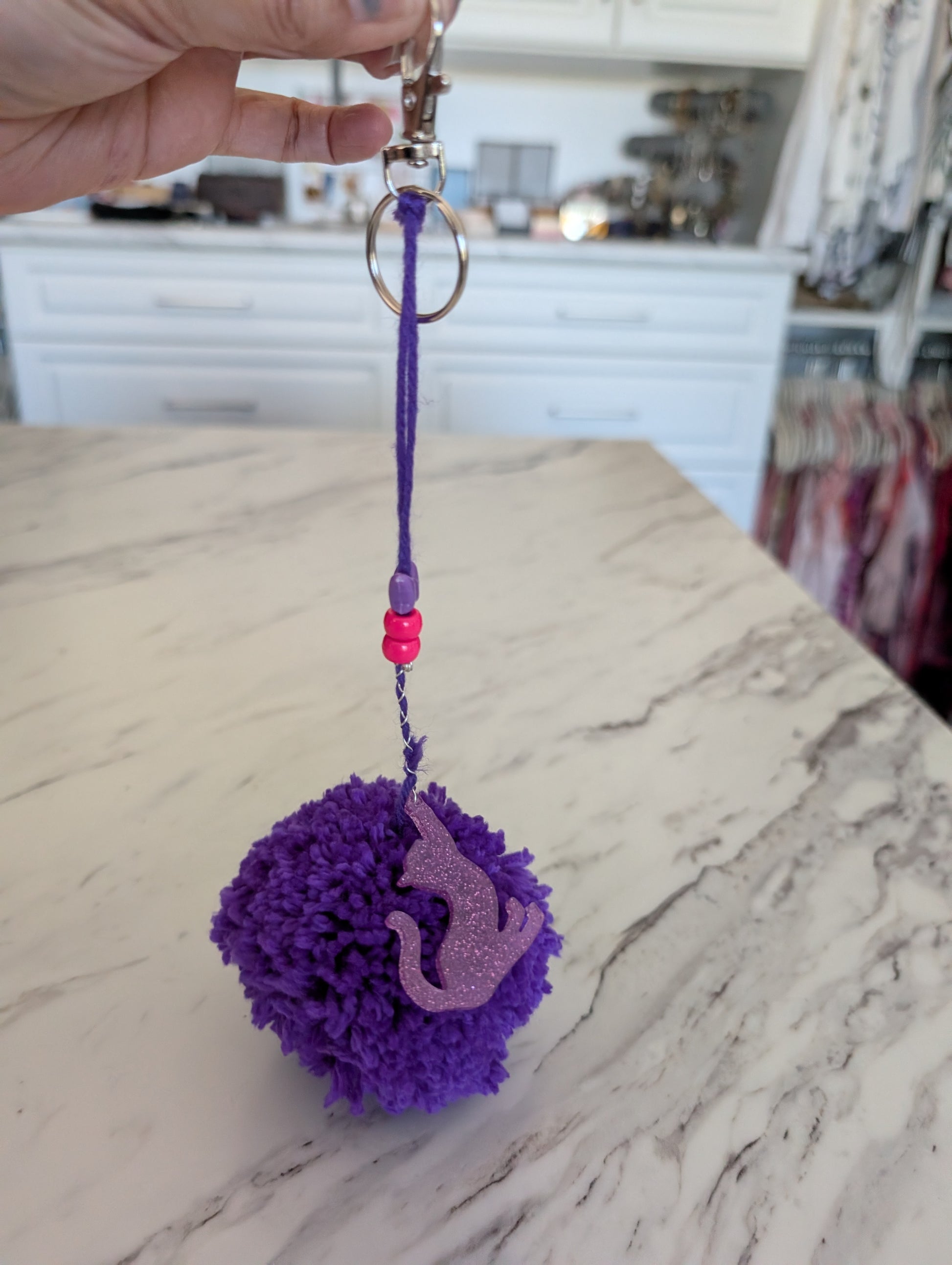 Purple pom-pom keychain with a cat charm on a marble countertop.