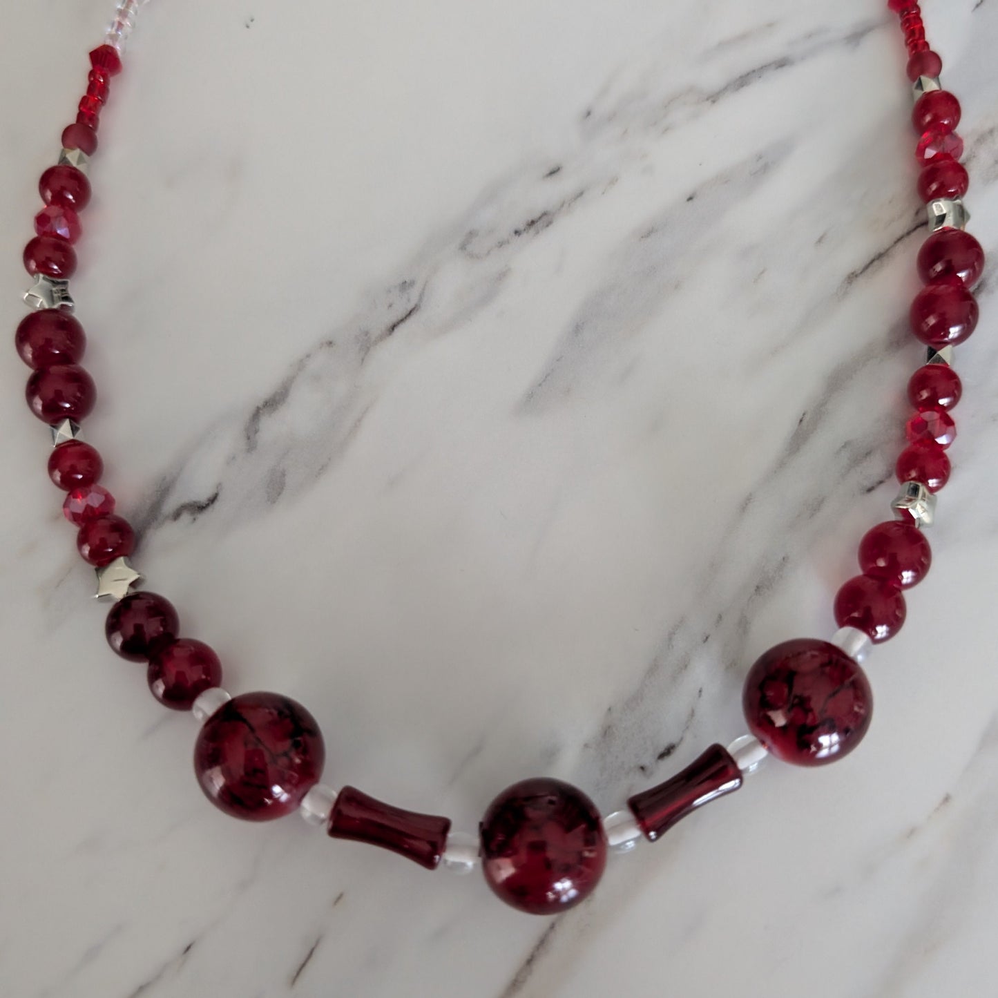 A beaded glass necklace with deep red marbled beads, chunky round beads, and small silver-toned spacer beads, displayed on a white marble surface.