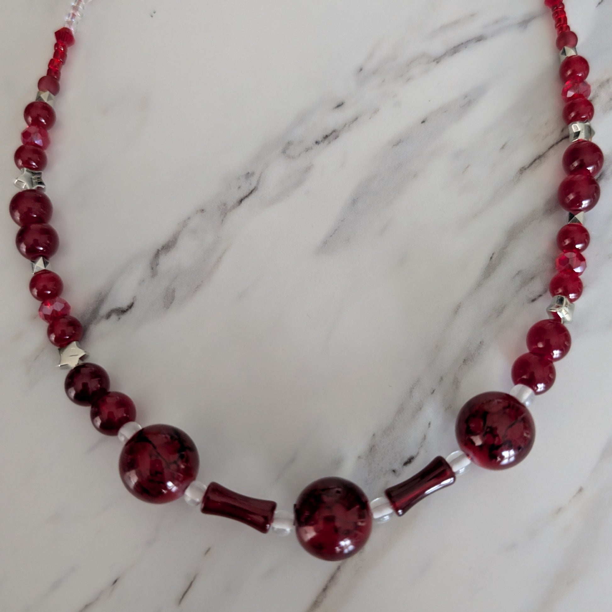 A beaded glass necklace with deep red marbled beads, chunky round beads, and small silver-toned spacer beads, displayed on a white marble surface.