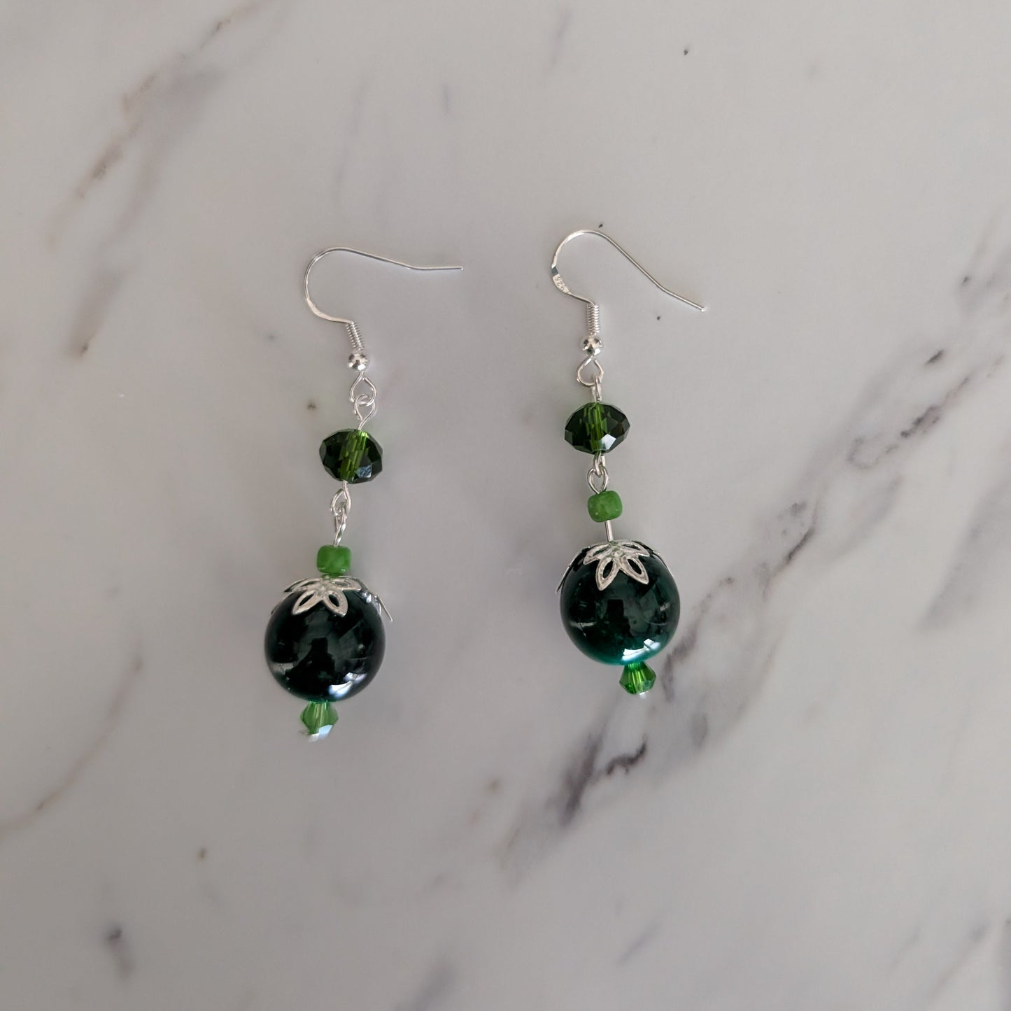 A pair of handmade dangle earrings with dark green faceted beads, small green accent beads, and silver fishhook wires, displayed on a white marble surface.