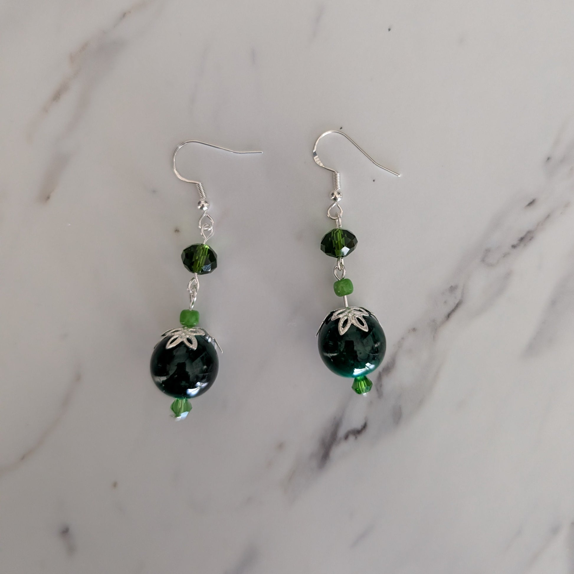 A pair of handmade dangle earrings with dark green faceted beads, small green accent beads, and silver fishhook wires, displayed on a white marble surface.