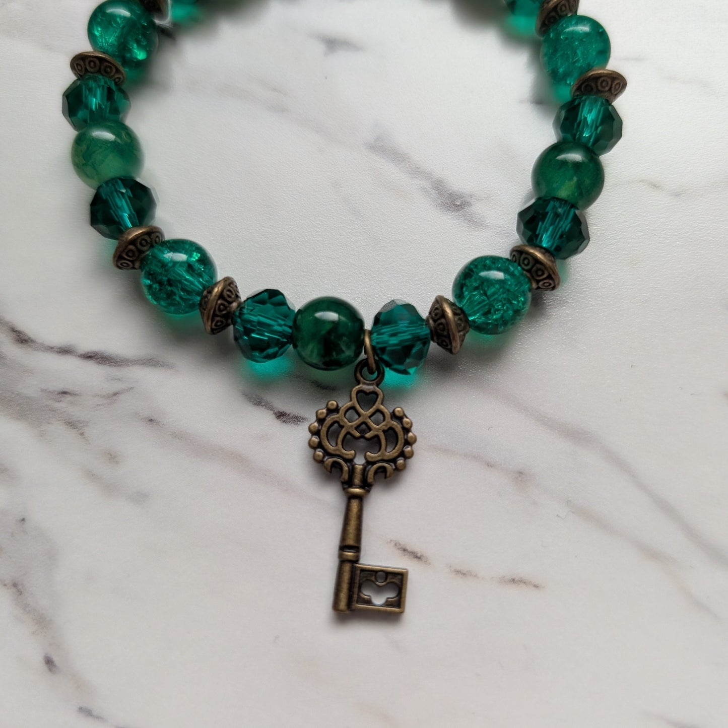 A beaded stretch bracelet featuring translucent green glass beads, small bronze spacer beads, and a dangling antique bronze key charm, displayed on a white marble surface.