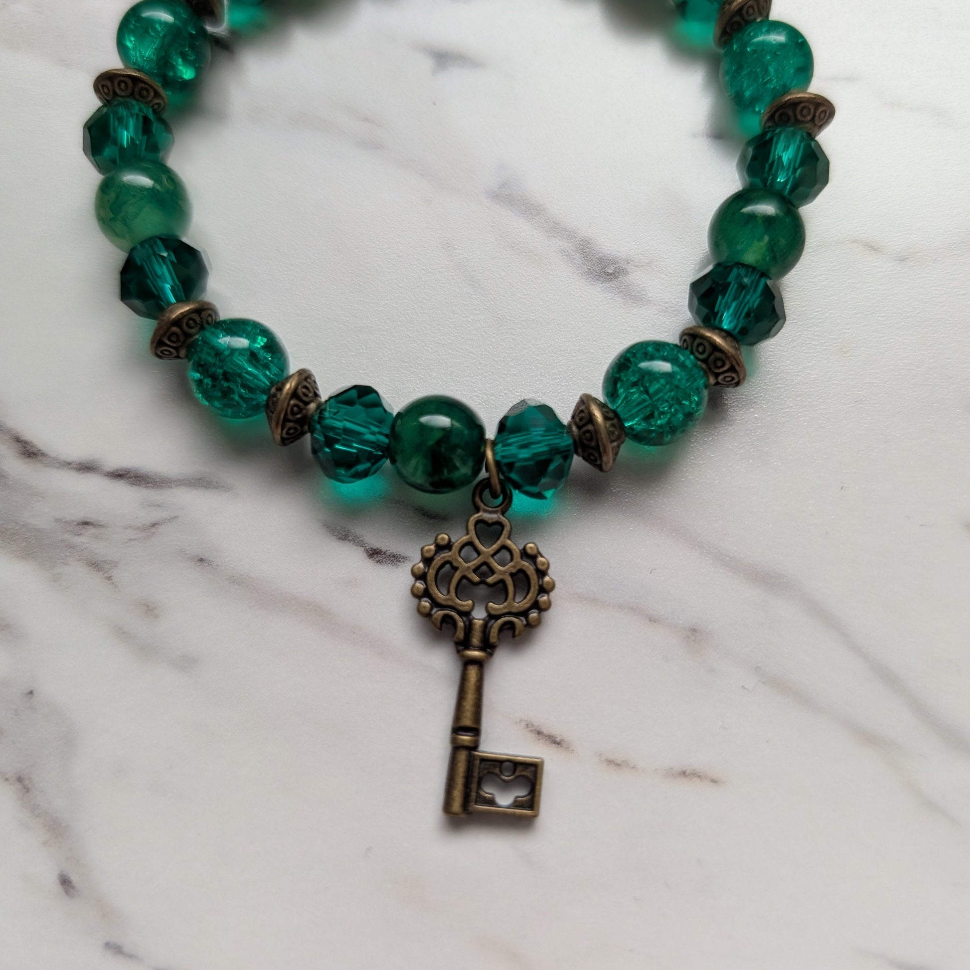 A beaded stretch bracelet featuring translucent green glass beads, small bronze spacer beads, and a dangling antique bronze key charm, displayed on a white marble surface.