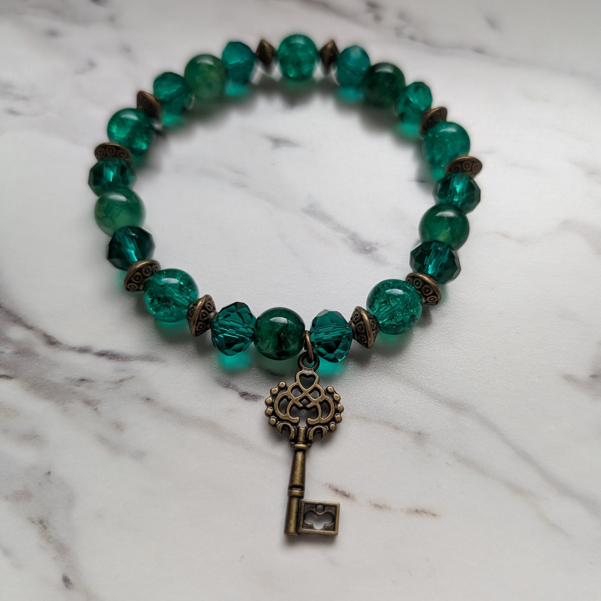 A beaded stretch bracelet featuring translucent green faceted beads, small bronze-toned spacer beads, and a dangling antique-style metal key charm, displayed on a white marble surface.