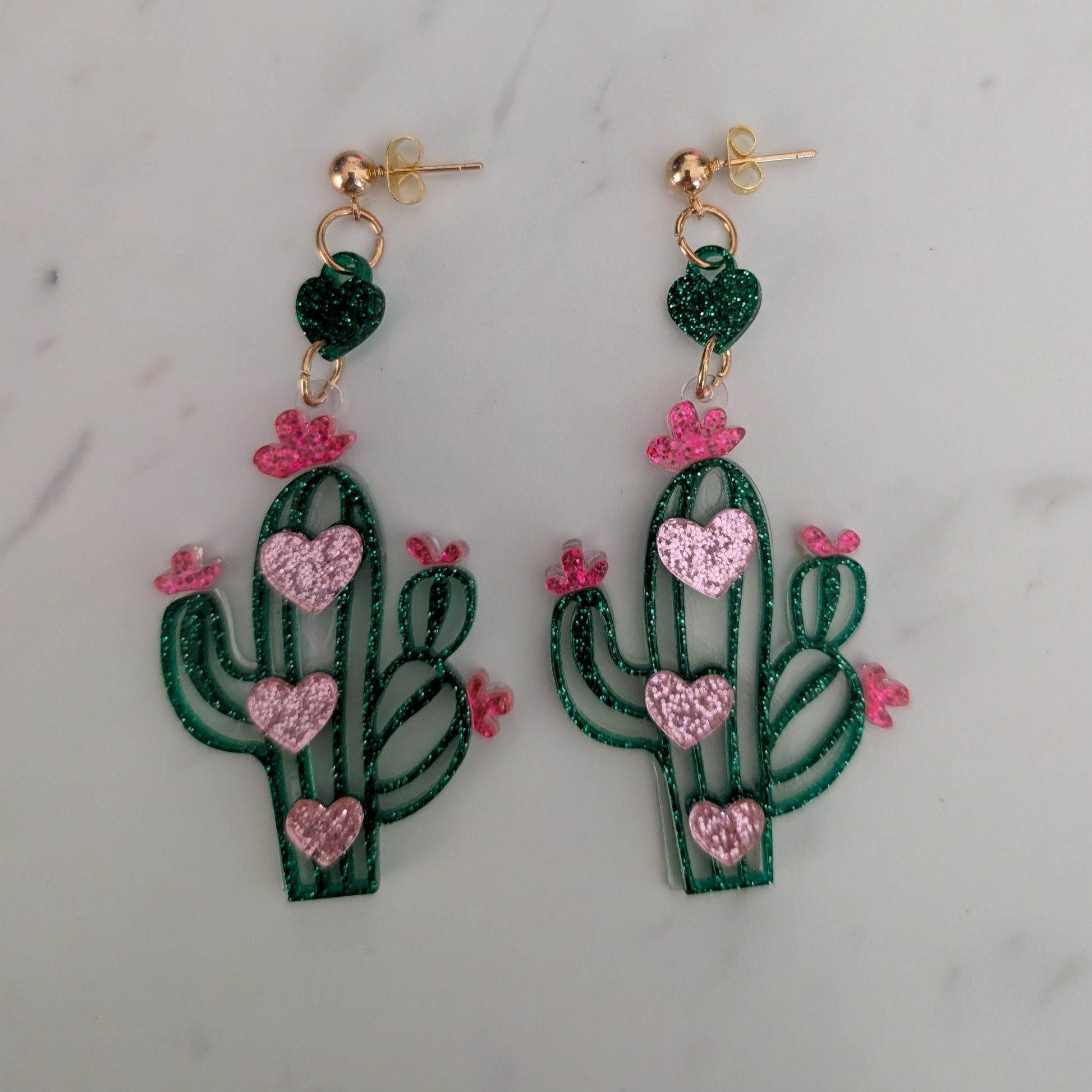 A pair of dangling cactus earrings with green glittery bodies, pink glittery heart-shaped details, and small pink flower accents, hanging from gold-tone posts on a light marble surface.