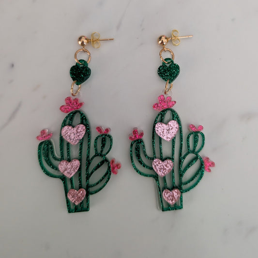 A pair of dangling cactus earrings with green glittery bodies, pink glittery heart-shaped details, and small pink flower accents, hanging from gold-tone posts on a light marble surface.