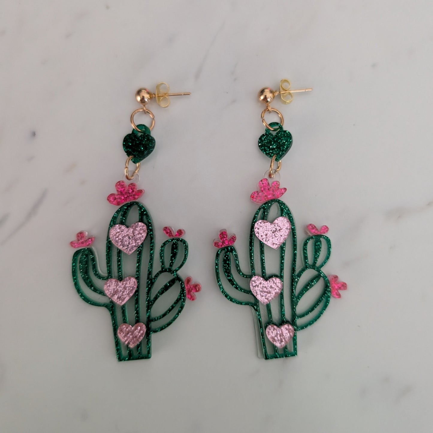 A pair of dangling cactus earrings with green glittery bodies, pink glittery hearts, and small pink flowers, hanging from gold-tone posts on a light marble surface.