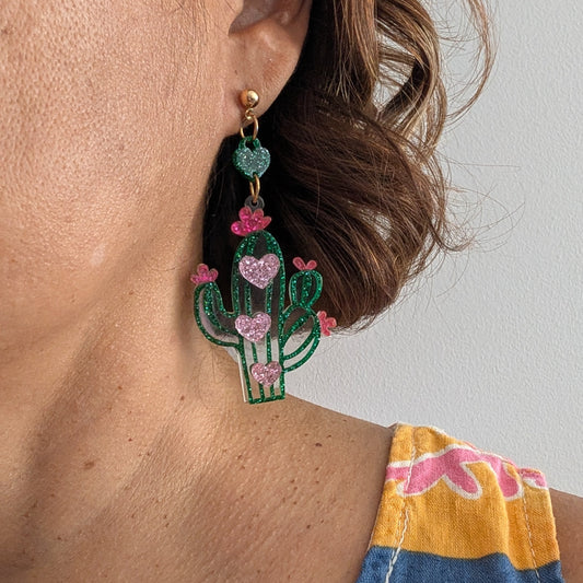 A close-up of a person wearing a dangling cactus earring with pink heart-shaped flowers and green textured details, paired with a colorful patterned top against a plain light gray wall.