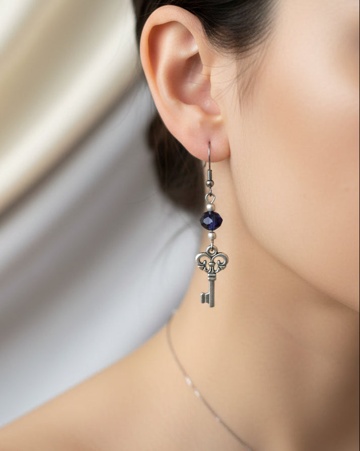 A pair of silver key dangle earrings with dark blue beads, displayed on a white marble surface.