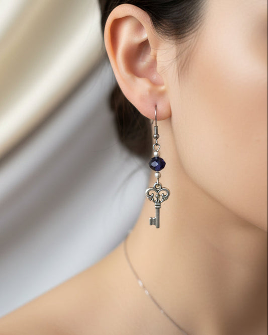 A pair of silver key dangle earrings with dark blue beads, displayed on a white marble surface.