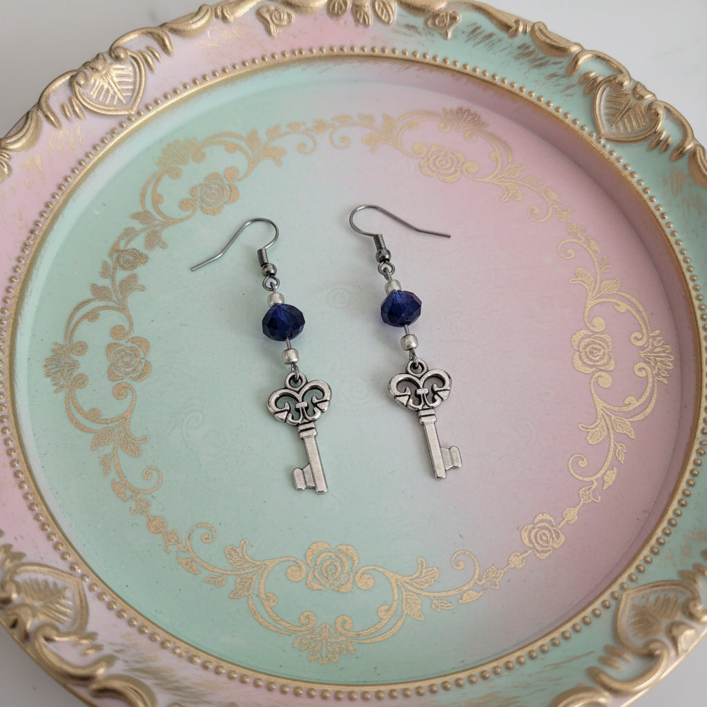 A pair of silver key dangle earrings with dark blue faceted beads, displayed on a white marble surface.