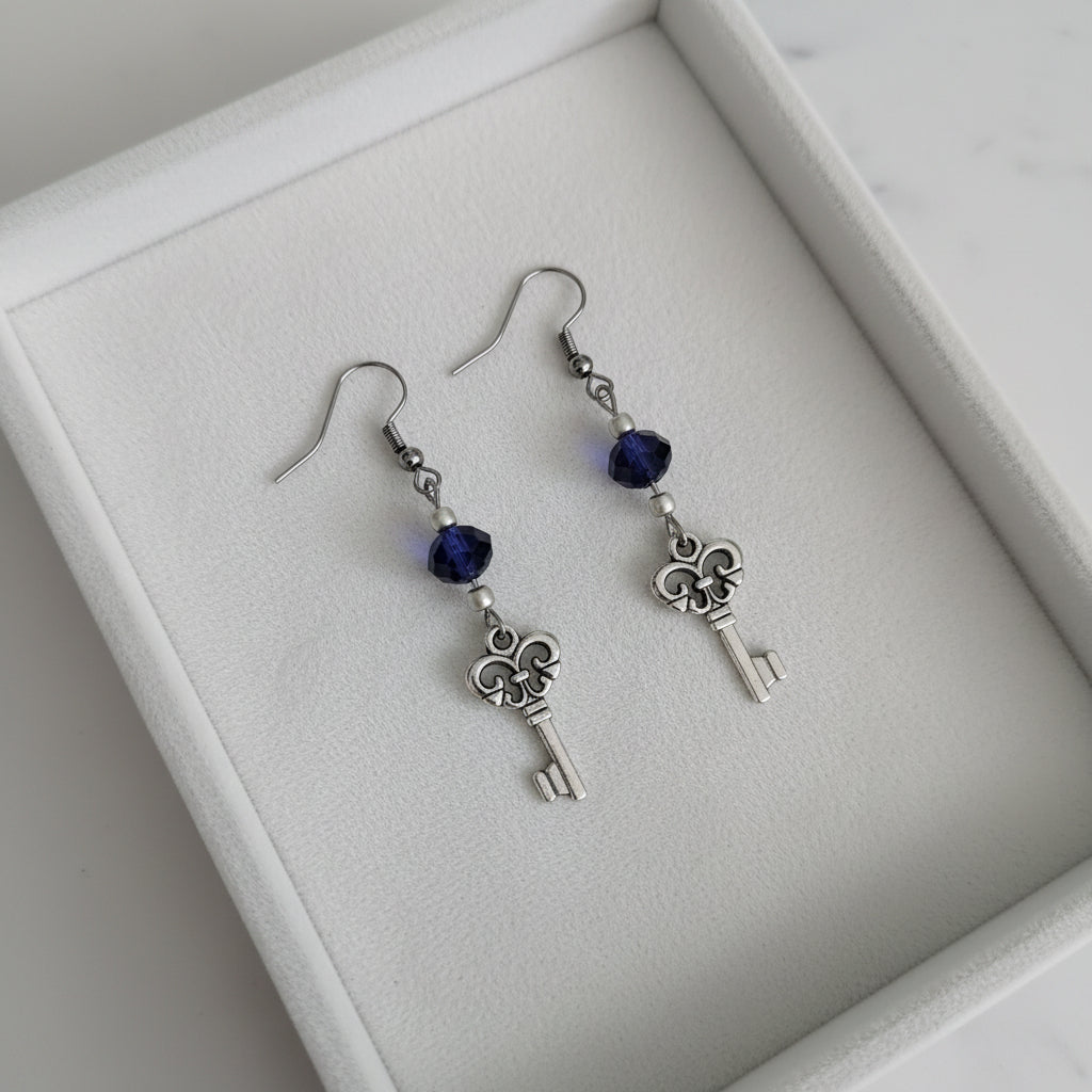A pair of silver key dangle earrings with dark blue faceted beads, displayed on a white marble surface.
