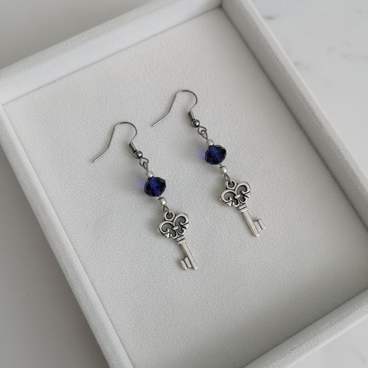 A pair of silver key dangle earrings with dark blue faceted beads, displayed on a white marble surface.