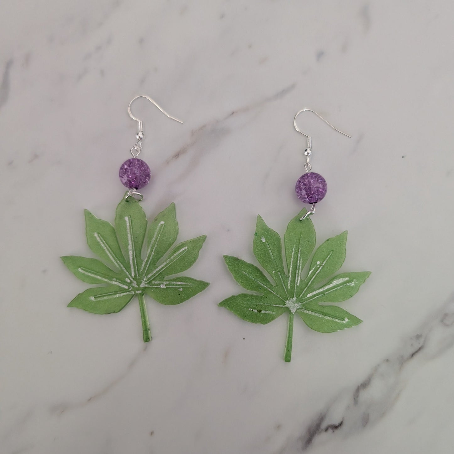 A pair of handmade dangle earrings featuring green maple leaf charms with white glitter accents, a small purple glitter bead, and silver fishhook wires, displayed on a white marble surface.