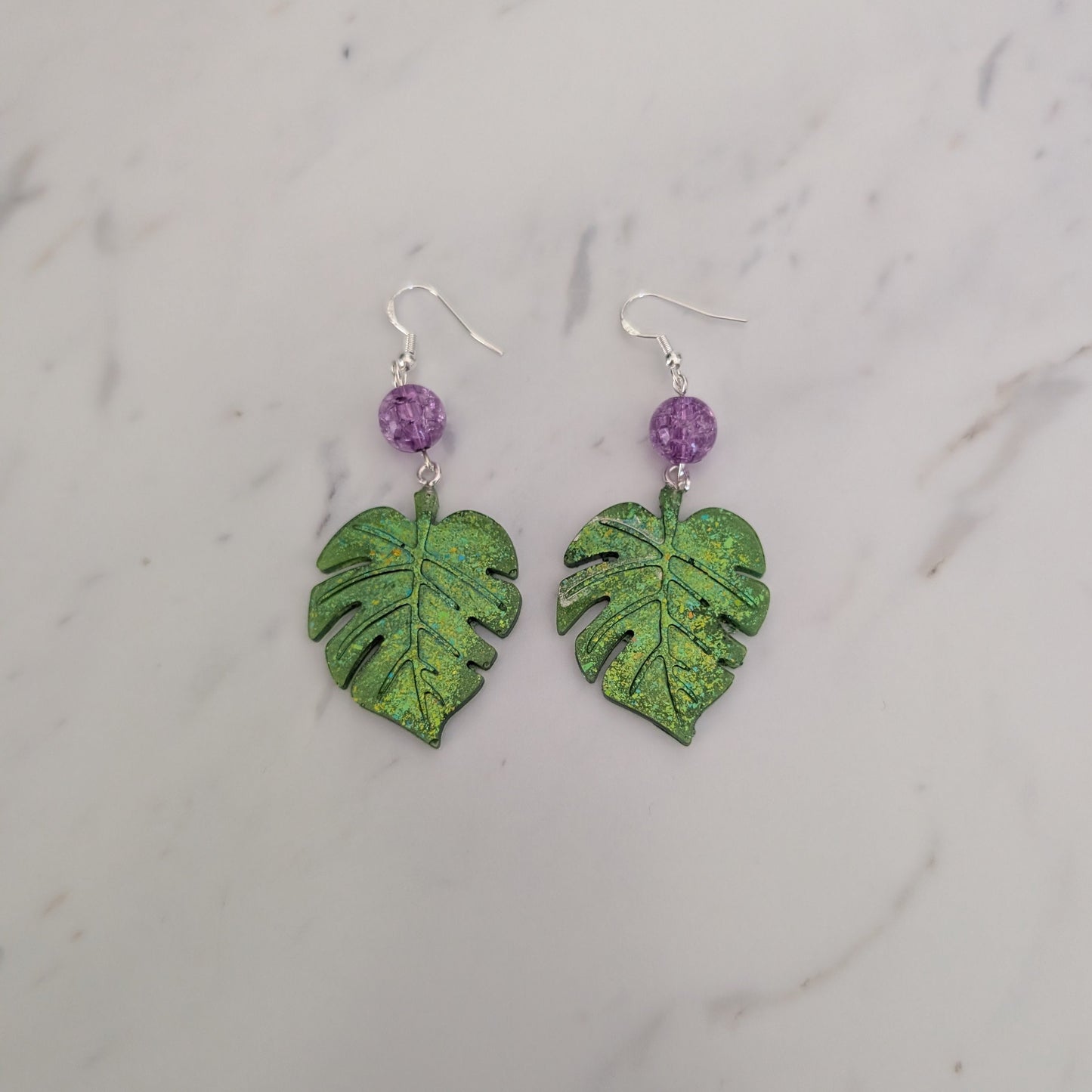 A pair of dangle earrings featuring textured green monstera leaf charms and small purple beads on silver hooks, displayed on a white marble surface.