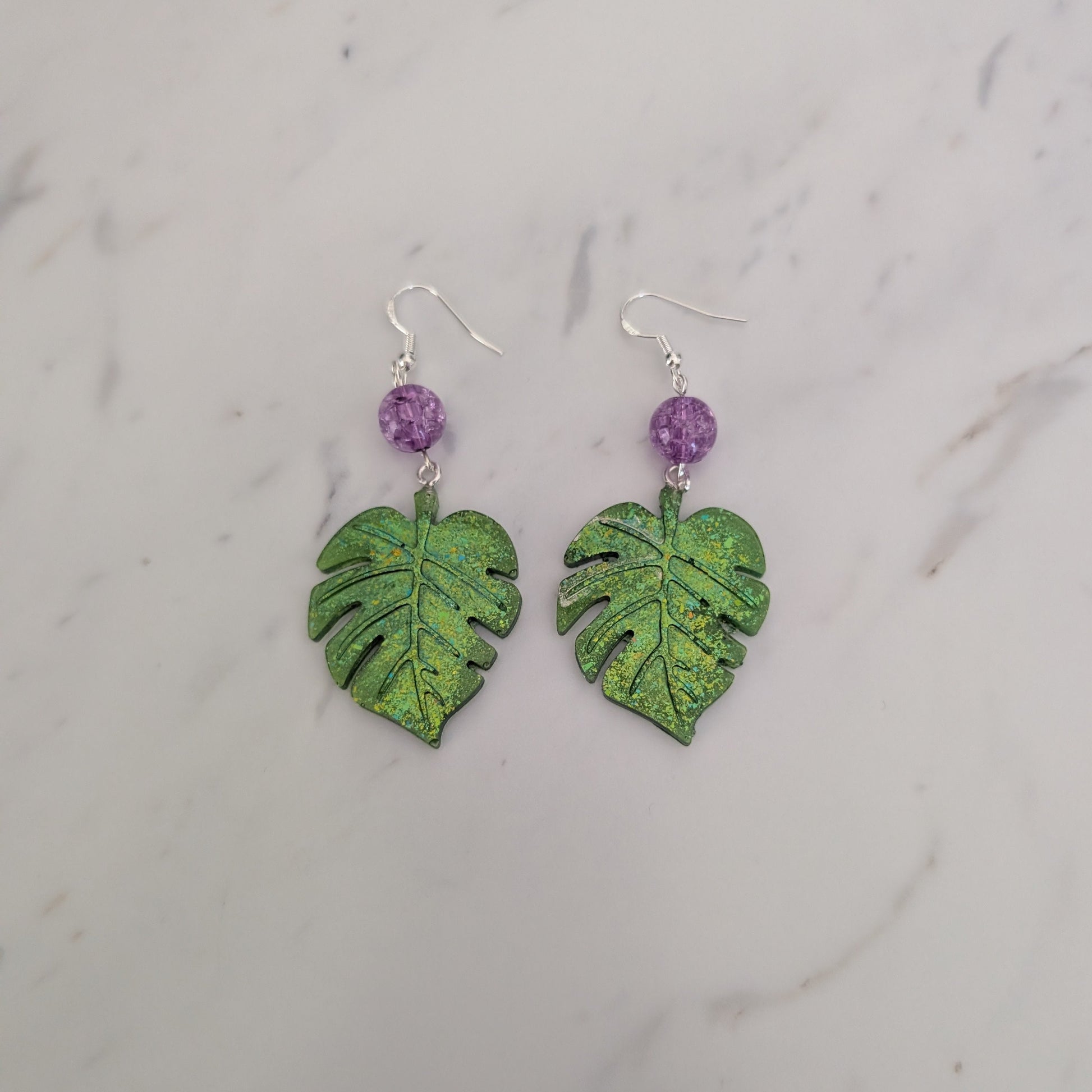 A pair of dangle earrings featuring textured green monstera leaf charms and small purple beads on silver hooks, displayed on a white marble surface.