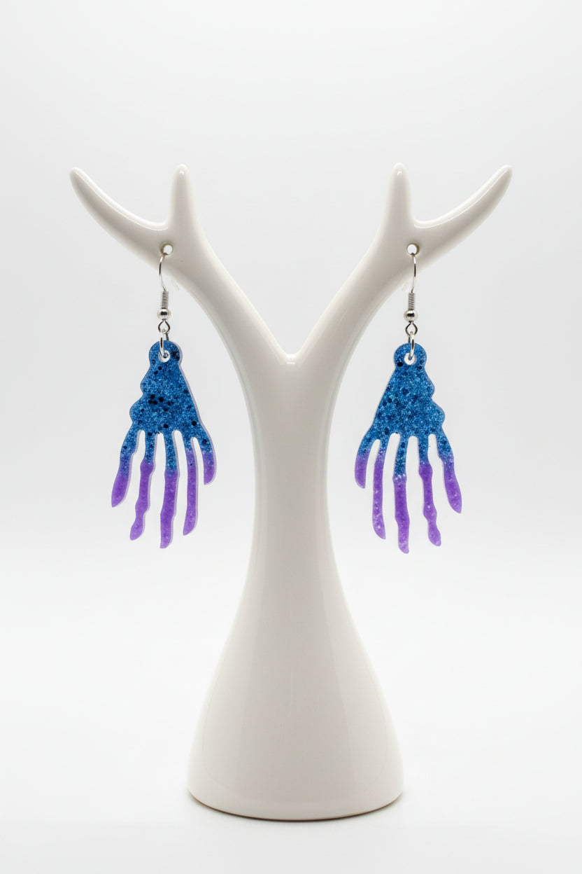 A pair of dangling earrings with glittery blue and purple hand-shaped charms on silver fishhook wires, displayed on a dark wood background.