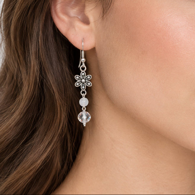 A close-up of a woman wearing a pair of silver dangle earrings with clear crystal-like beads and floral details.