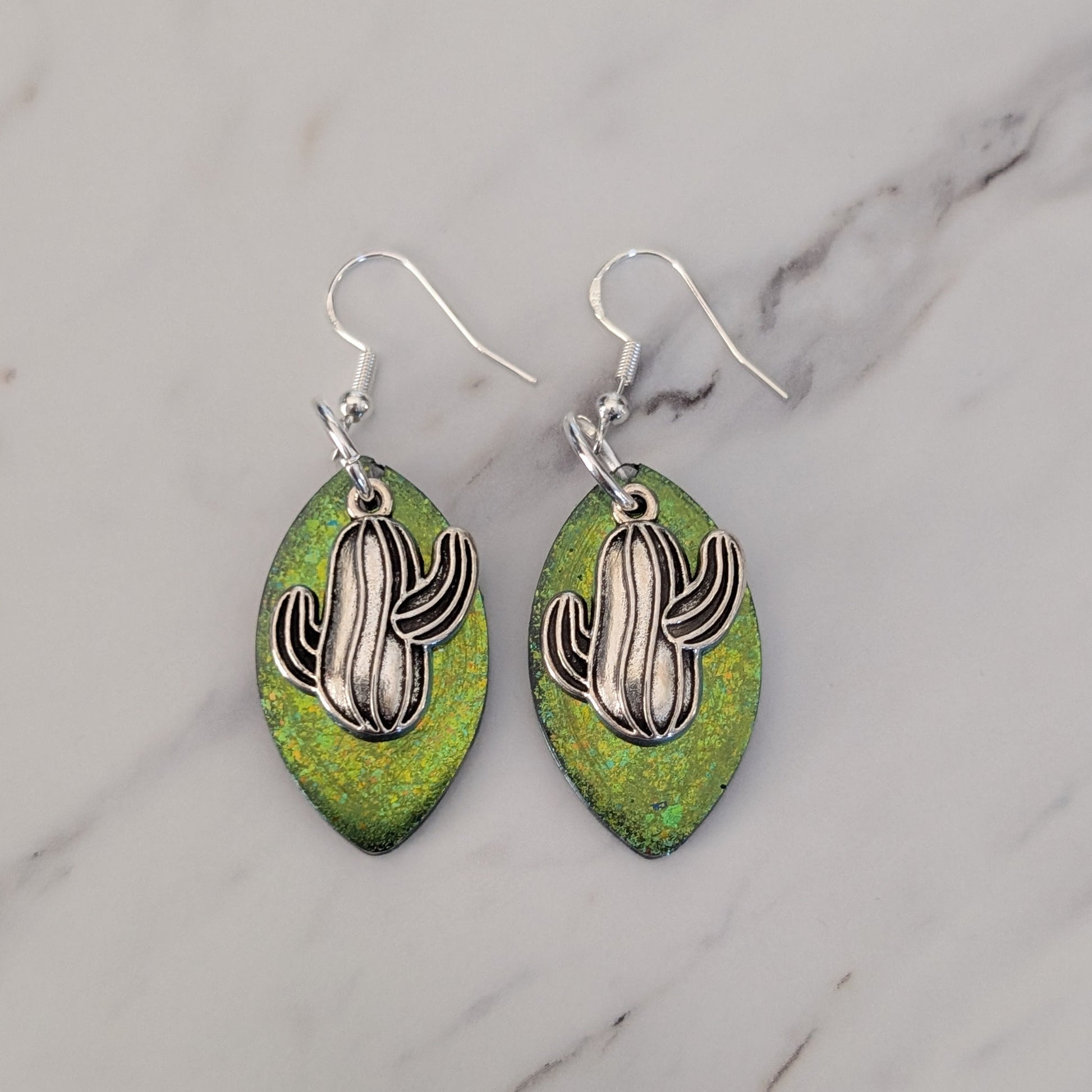 Green leaf-shaped earrings with silver cactus designs on a marble background