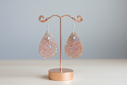 Teardrop-shaped glittery earrings with starburst designs on a pink background