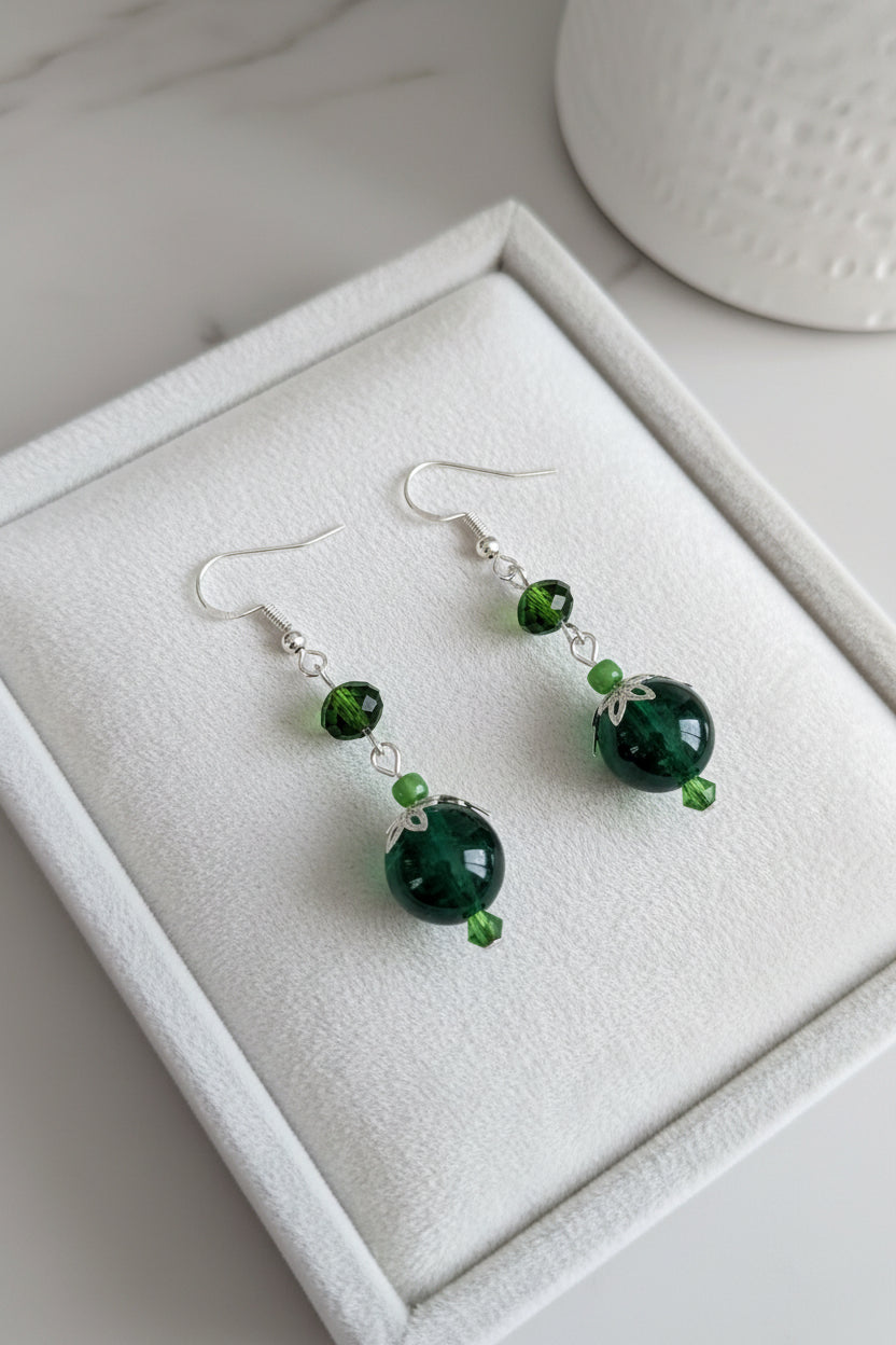A pair of green beaded dangle earrings with silver fishhook wires, displayed on a white textured jewelry box.