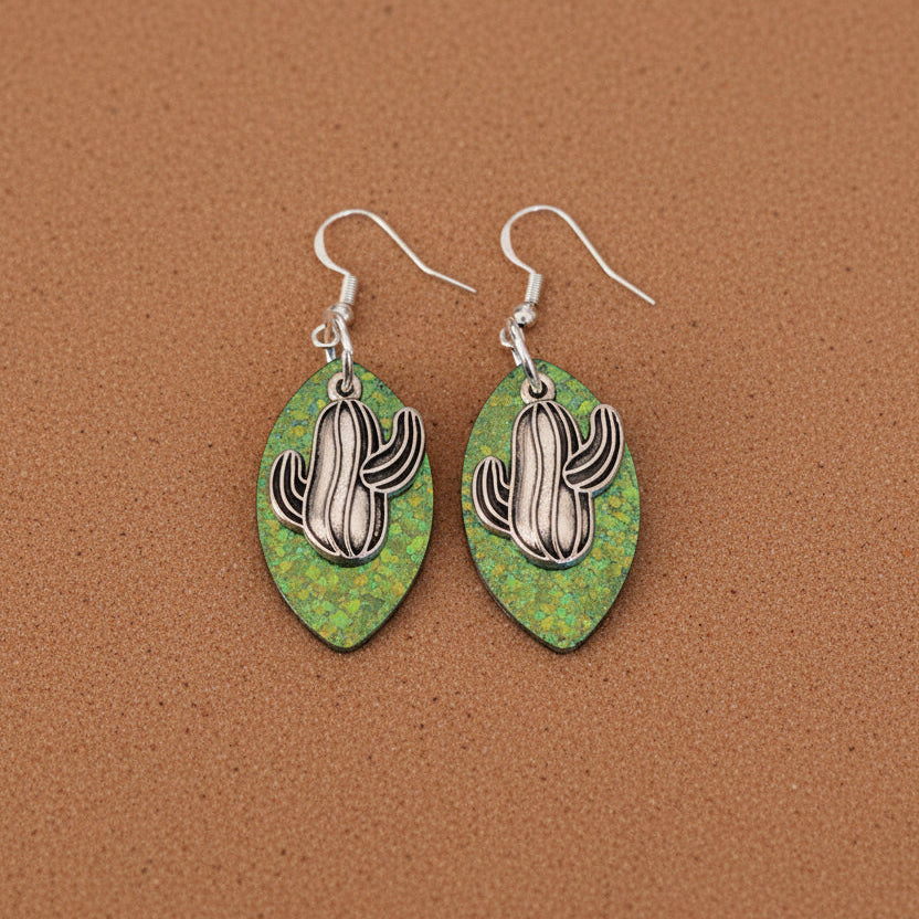 Pair of earrings with cactus design on green resin leaf shape against a brown background 