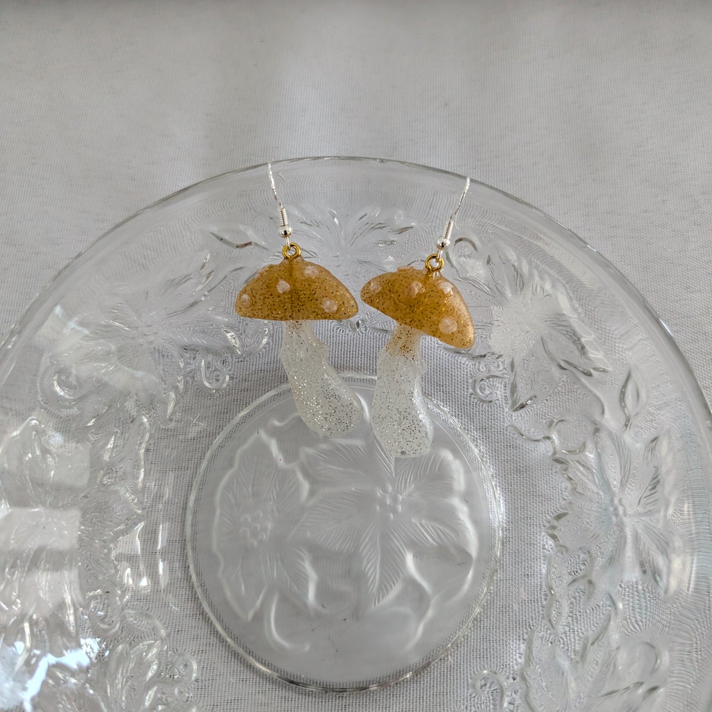 Mushroom-shaped earrings on a decorative glass plate with a white cloth background