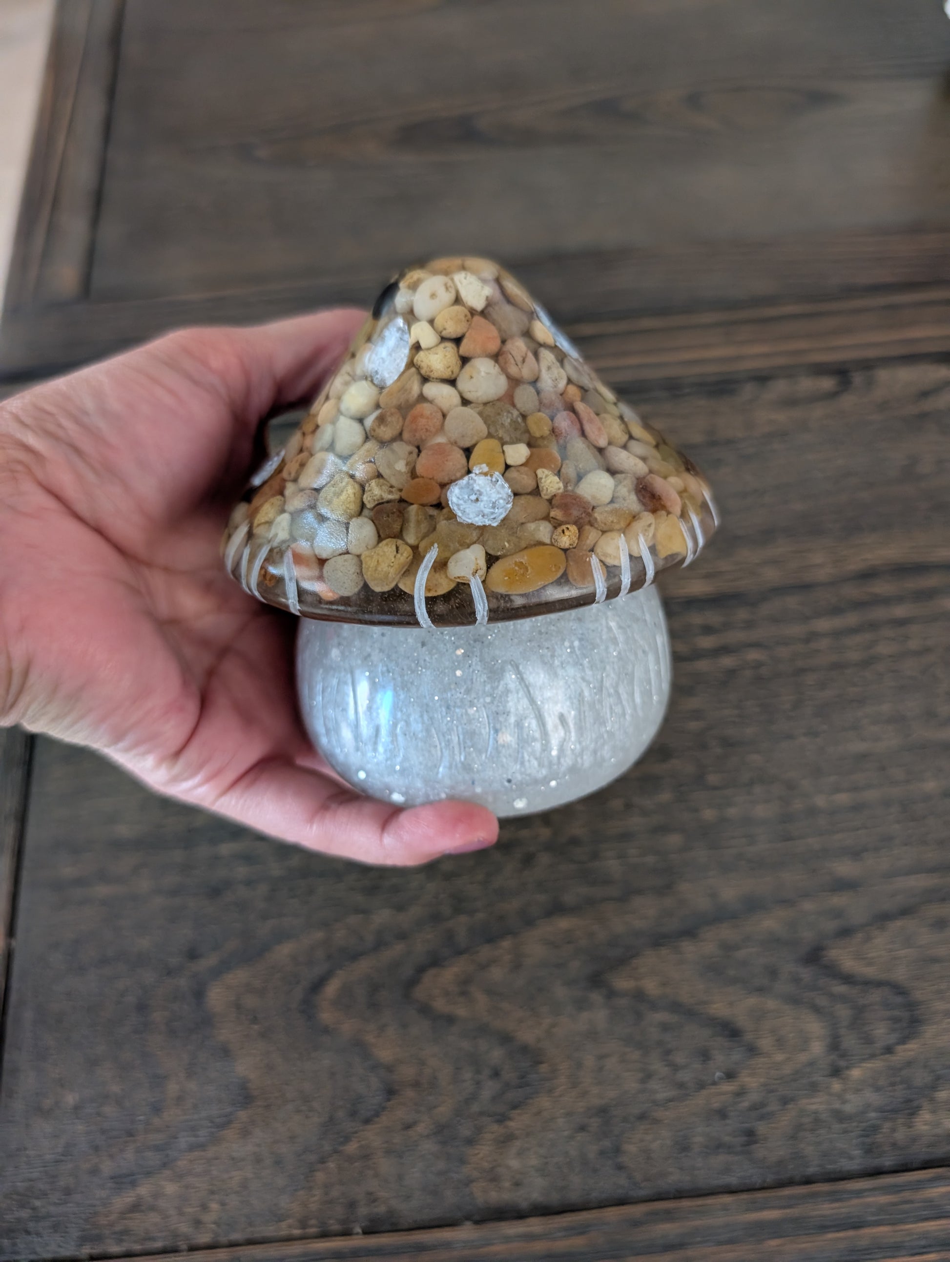 Hand holding a decorative mushroom trinket with lid on a wooden table.